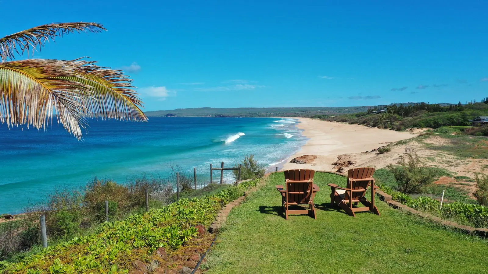 Two chairs on a grassy bluff overlooking a long white-sand beach on Molokaʻi’s West End